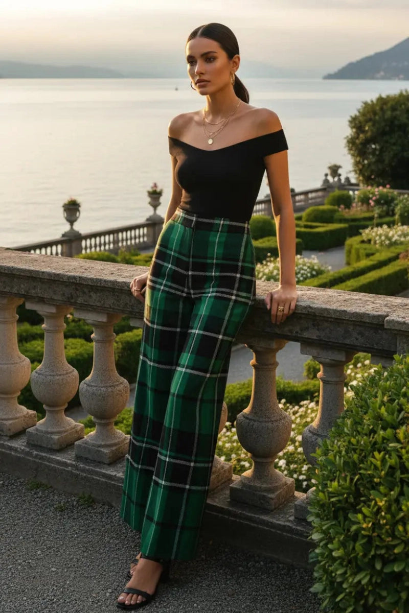 Woman wearing green tartan wide leg trousers and black off-shoulder top standing by stone railing