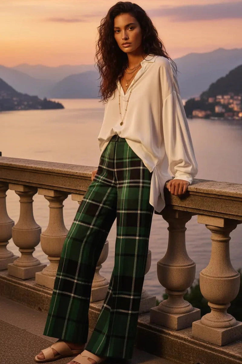 Woman wearing green tartan wide leg trousers paired with a white blouse standing by a scenic waterfront balcony
